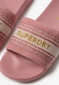 Sandalias de slide rosas con una suela de goma texturizada, tira trenzada con patrones en rosa y blanco, y "SUPERDRY" bordado en dorado.