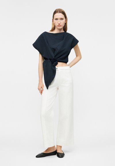 Γυναίκα που φορεί μαύρο τοπ με κοντά μανίκια και μεγάλο δέσιμό στο πλάι, λευκό παντελόνι με φαρδιά μπατζάκια και μαύρα μπαλαρίνια, στέκεται μπροστά σε απλό φόντο.