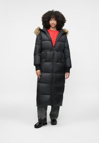 Zwarte pufferjas met een met bont gevoerd capuchon, lange uitvoering, ritssluiting en twee voorzakken. Gedragen over een rode top en gecombineerd met donkere broek.