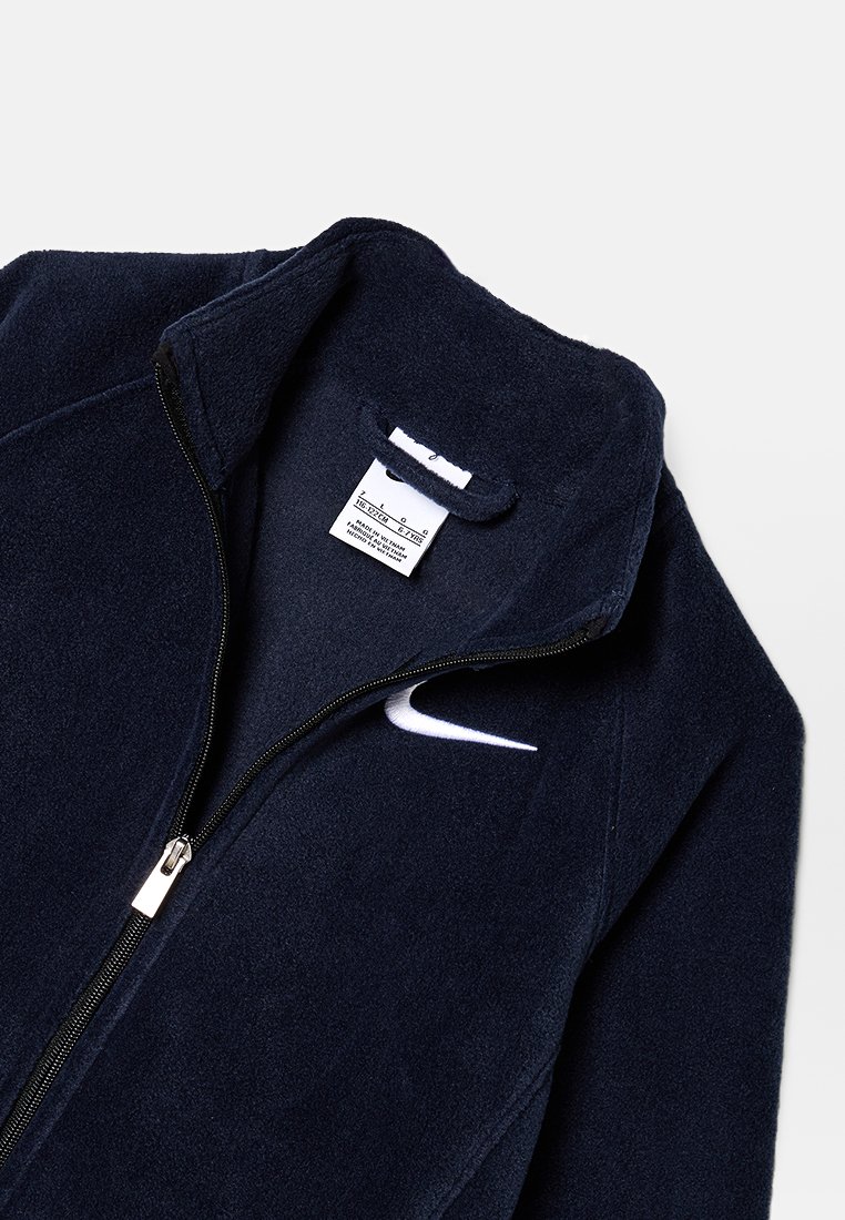 Veste en polaire bleu marine avec un col montant, fermeture éclair à l'avant, logo Nike blanc et étiquette d'entretien visible à l'intérieur de la zone du col.