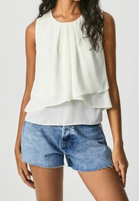 Ærmeløs lagdelt bluse i lys creme med plisseringer, parret med slidte denimshorts. Glat stof, afslappet pasform og blød tekstur.