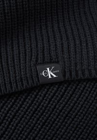 Fekete kötött szövet bordás textúrával, egy téglalap alakú fekete címkével, amelyen a "Calvin Klein Jeans" logo található.