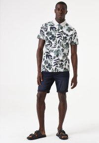 Korte mouwen bloesemshirt in het wit met groene en zwarte patronen, gecombineerd met donkere spijkerbroekshorts. Zwarte sandalen maken de outfit compleet.