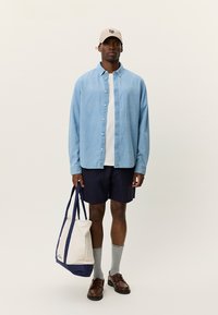 Ανοιχτό μπλε πουκάμισο με κουμπιά πάνω από λευκό t-shirt, σκούρο μπλε σορτς, γκρι κάλτσες, καφέ παπούτσια και μπεζ καπέλο. Κρατιέται μια υφασμάτινη τσάντα με μπλε λεπτομέρειες.