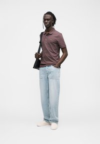 Braunes Polohemd mit kleinem goldenem Logo, hellblaue Jeans mit weitem Bein und beigen Schuhen. Das Modell trägt eine schwarze Schultertasche und hat geflochtenes Haar.