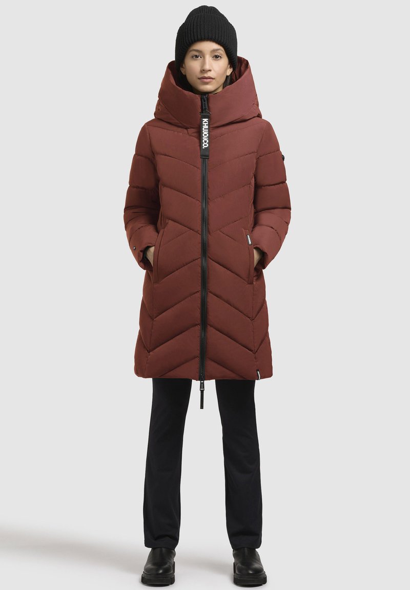 Cappotto imbottito marrone con motivo a zig zag, colletto alto e cappuccio. Presenta una chiusura frontale con zip e tasche laterali. Abbinato a pantaloni neri e stivali.