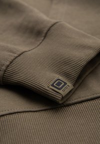 Gros plan du poignet en tissu côtelé marron d’un sweat-shirt avec une petite étiquette de marque carrée cousue près de la couture.