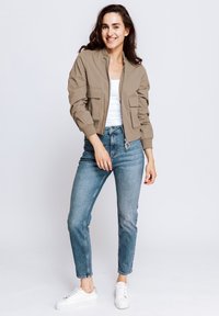 Khaki bomber jacket with two front pockets, paired with light blue jeans. White sneakers complete the outfit. Soft texture with casual design.