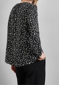 Svart topp med vitt abstrakt mönster, långa ärmar, rund halsringning och lös passform. Tyget verkar slätt med minimal textural detalj.