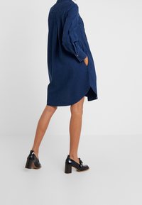 Džínová košilová šaty v tmavě modré barvě, s volným střihem, zapínáním na knoflíky, dlouhými rukávy a bočními kapsami. Kombinováno s černými lodičkami.