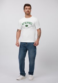 Hvid kortærmet T-shirt med grøn tekst "NEWPORT 1957 Sporting Goods", parret med blå jeans og hvide sneakers. Enkel design, afslappet pasform.