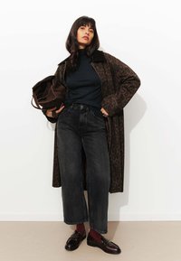 Manteau long à imprimé léopard brun sur un pull bleu marine, associé à un jean noir à jambes larges et des mocassins polis. Femme tenant un sac fourre-tout marron.