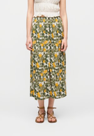 Donna che indossa una gonna midi con bottoni sul davanti e stampa floreale gialla, abbinata a sandali marroni con cinturini e una canotta bianca senza maniche.