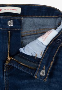 Ciemnoniebieskie ultraskinny jeansy z metalowym zamkiem błyskawicznym, zapięciem na guzik oraz kontrastowym złotym przeszyciem. Podszewka widoczna wewnątrz w pasie.