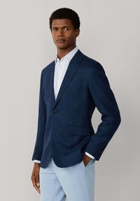 Mann trägt marineblauen Blazer, weiß gestreiftes Hemd und hellblaue Hose und steht mit einer Hand in der Tasche vor einem einfarbigen Hintergrund.
