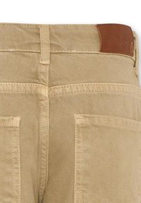 Pantaloni beige con una texture morbida, dotati di un'etichetta in pelle sul cinturino e due ampie tasche a toppa sul retro.