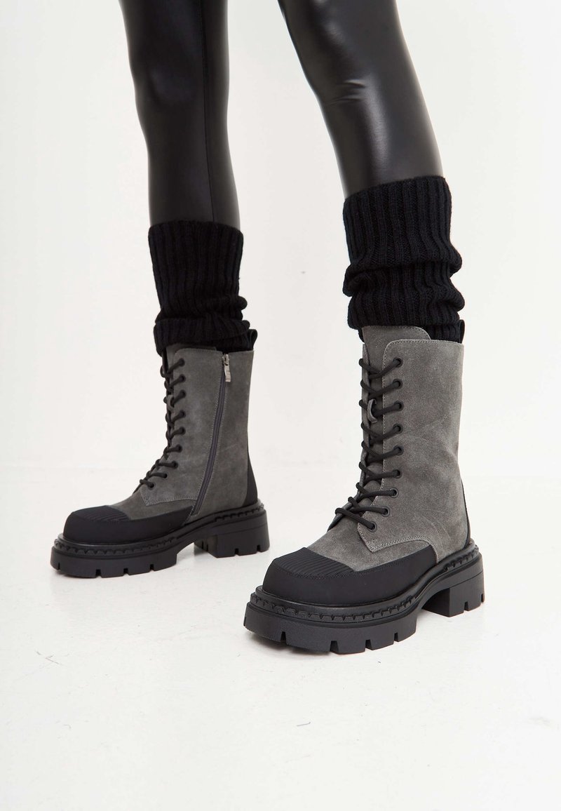 Grijze suède veterschoenen met een zwarte rubberen zool, voorzien van een zijrits, dikke zool en geribbelde zwarte gebreide beenwarmers.