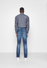 Ciemnoniebieskie jeansy o dopasowanym kroju, z detalami przetarć, oraz białe trampki. Model ma na sobie szarą koszulę zapinaną na guziki z teksturowanym wzorem.