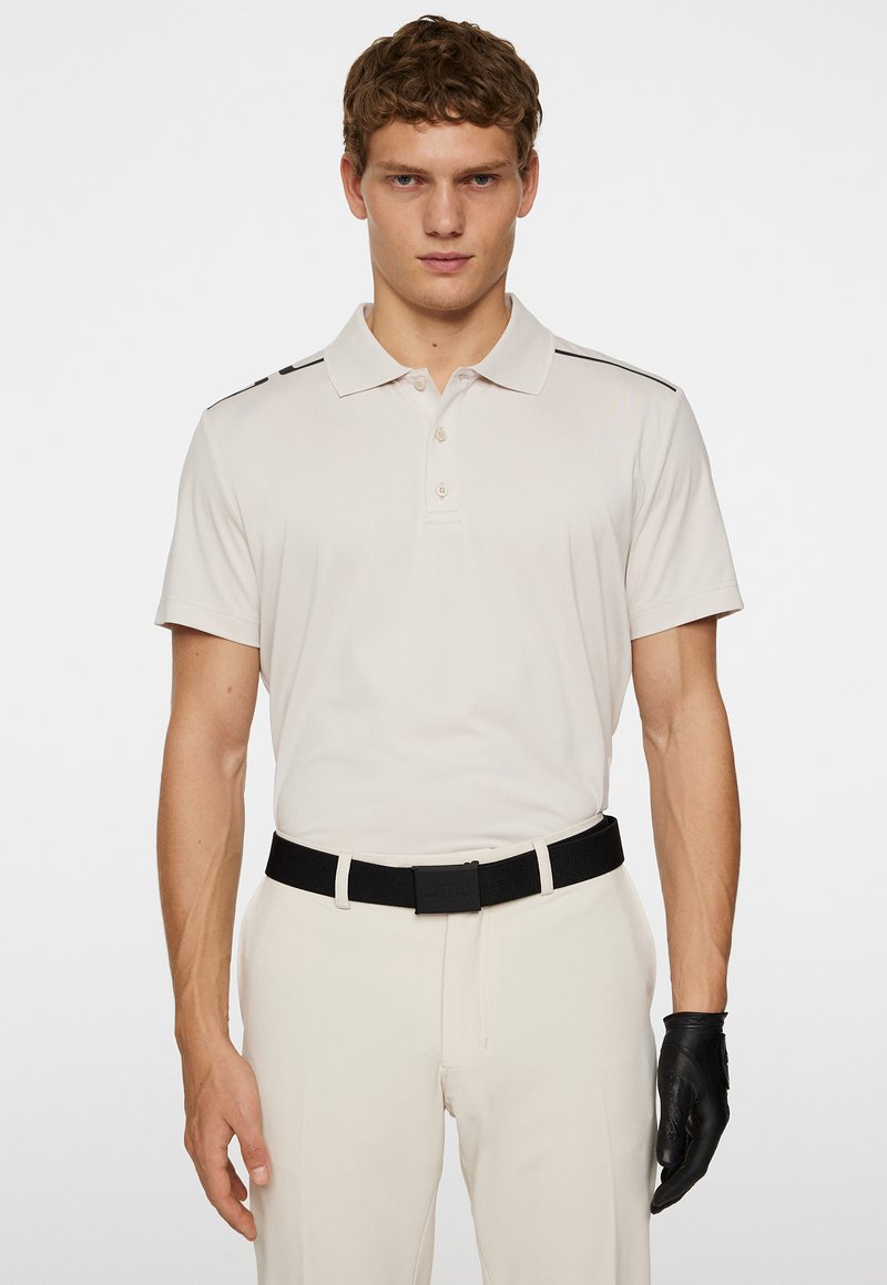 Beige kortärmad polo skjorta med krage, med svarta axelränder. Matchad med ljusa byxor och ett svart bälte.