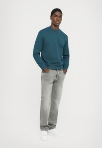 Teal sweatshirt med rund halsringning, kombinerat med ljusgrå jeans. Modellen bär vita sneakers. Enkelt, utan utsmyckningar design.