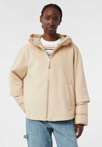 Giacca beige con zip e cappuccio, caratterizzata da una texture morbida e maniche larghe, indossata sopra un dolcevita a righe. I pantaloni in denim blu sono visibili.