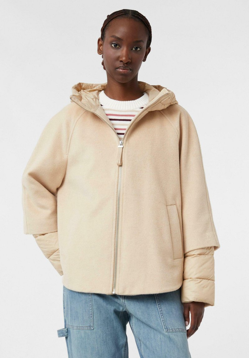 Giacca beige con zip e cappuccio, caratterizzata da una texture morbida e maniche larghe, indossata sopra un dolcevita a righe. I pantaloni in denim blu sono visibili.