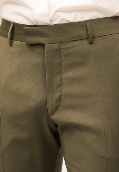 Παντελόνι σε ελιά πράσινο με κρυφό μπροστινό φερμουάρ και θηλιές για ζώνη, φορεμένο με ένα άσπρο πουκάμισο μπαλαρισμένο μέσα.