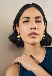 Gold- und blaue Tropfenohringe mit runden Perlen; glatte Textur, geschichtetes Design mit einer Mischung aus Materialien und Formen. Das Modell hat kurze Haare.
