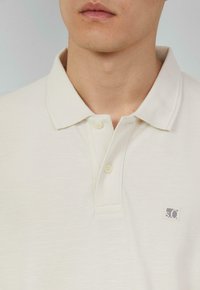 Polo en beige clair, fabriqué à partir d'un tissu texturé. Il présente un col classique, trois boutons et un petit logo sur la poitrine gauche.