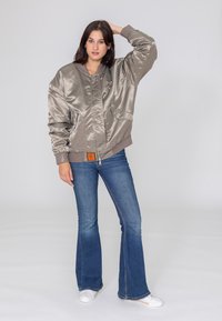 Khaki satynowa kurtka bomberka z przednimi kieszeniami, ściągaczami przy mankietach i zamek błyskawiczny, w zestawieniu z dżinsami w stylu dzwonów w kolorze niebieskim oraz białymi butami typu slip-on.