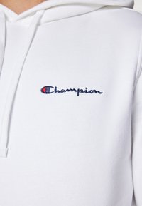 Fehér kapucnis pulóver puha anyagból, kék és piros hímzett "Champion" logóval a mellkason. Egyszerű, hétköznapi design.