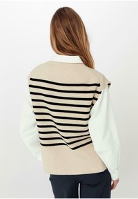 Béžový pletený svetr s černými horizontálními pruhy. Má spadlá ramena a vroubkovaný lem. Nosič má na sobě světle modrou košili s knoflíky.