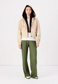 Veste à capuche beige courte avec des poignets élastiques, assortie à un pantalon large plissé vert et des chaussures blanches à bout pointu.