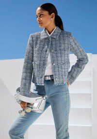 Blauwe tweed jas met kraag en franjes aan de onderkant, gecombineerd met lichte spijkerbroeken met decoratieve zijaccenten. Zilveren handtas en een zonnebril.