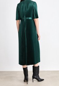 Groene plooienjurk met korte mouwen, satijnen riem en een watervalontwerp, gecombineerd met zwarte enkellaarsjes met punten en hakken.