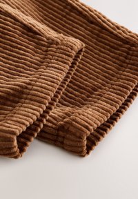 Tissu en velours côtelé marron avec de larges côtes verticales, plié soigneusement sur une surface blanche et couture visible le long du bord.