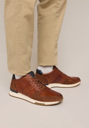 Bruine leren en suède sneakers met witte zolen gedragen met beige broek en witte sokken op een lichte achtergrond.