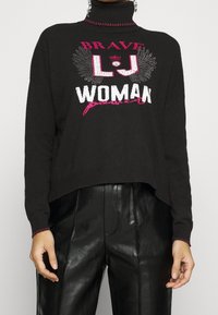 Pull court noir avec un col montant, arborant un texte graphique rose et blanc "BRAVE LJ WOMAN" et des accents décoratifs le long du col.