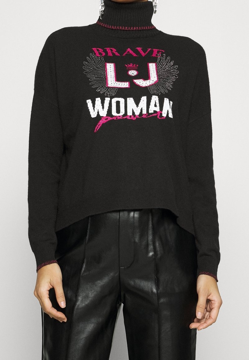 Pull court noir avec un col montant, arborant un texte graphique rose et blanc "BRAVE LJ WOMAN" et des accents décoratifs le long du col.
