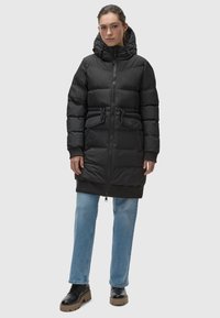 Black puffer coat with a hood, featuring a front zipper, drawstring waist, and two front pockets, paired with light blue jeans and black shoes.