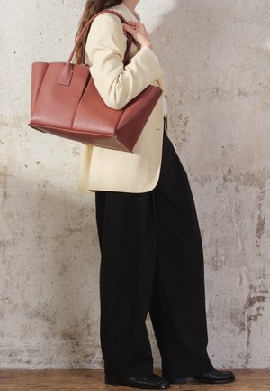 Borsa tote in pelle marrone con lati strutturati e doppi manici, esposta su uno sfondo di cemento, caratterizzata da un design minimalista e una texture liscia.