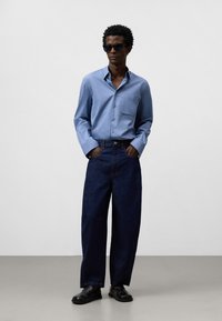 Ljusblå knapp-ned skjorta med bröstficka, kombinerad med mörkblå vida jeans och svarta slip-on skor, mot en neutral bakgrund.