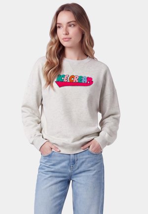 Giovane donna con capelli lunghi e mossi che indossa una felpa beige chiaro con testo colorato "ICE CREAM" e jeans azzurri, mani nelle tasche.