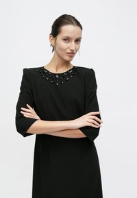 Μαύρο φόρεμα με κοντά μανίκια, με λαιμόκοψη διακοσμημένη με χάντρες. Το ύφασμα φαίνεται λείο και δομημένο, με ίσια σιλουέτα.