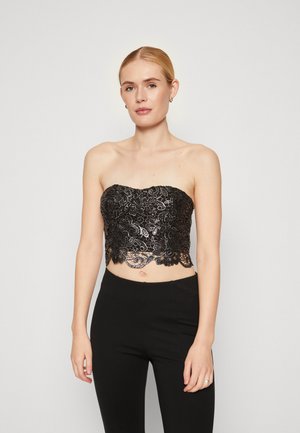 Melnas mežģīņu strapless topiņš ar sarežģītiem ziedu rakstiem un viļņotu apakšmalu, apvienots ar melniem pielāgotajiem biksēm.