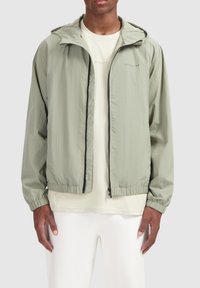 Lichtgroene windbreaker jas met een capuchon, enkele voorkant rits, elastische manchetten en zoom. Draagt over een beige shirt, minimalistisch ontwerp.