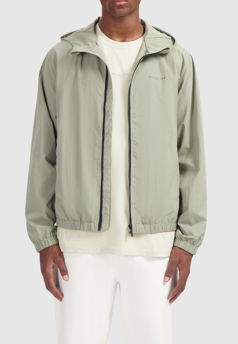 Lichtgroene windbreaker jas met een capuchon, enkele voorkant rits, elastische manchetten en zoom. Draagt over een beige shirt, minimalistisch ontwerp.