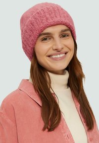 Un bonnet rose en tricot avec une texture douce, présentant un motif à côtes. Porté avec un pull à col roulé crème clair et une chemise en velours côtelé rose.