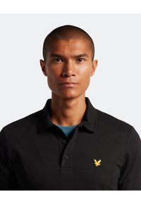 Svart piké med krage, prydd med en gul örn-emblem på vänster bröst. Slät textur och korta ärmar.