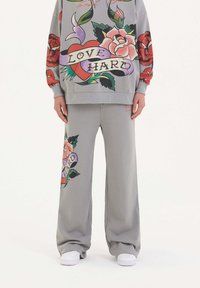 Grå sweatshirt och matchande byxor med färgglada tatuering-liknande mönster, inklusive rosor och banderoller med texten "ÄLSKA HÅRT". Mjuk tyg, lös passform.